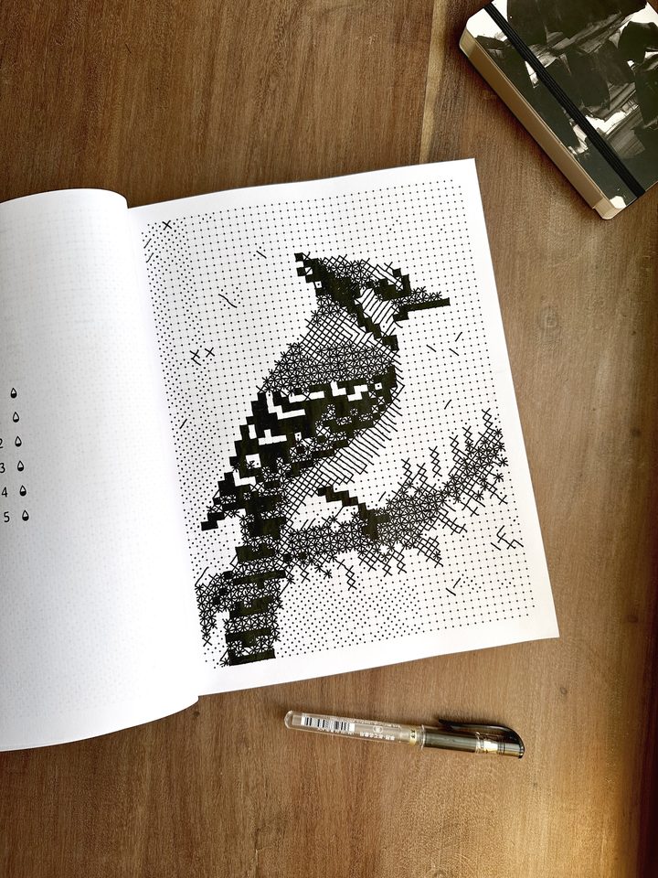 The monochrome book open on a finished page, a blue jay on a branch drawn in black marks, the pen lying below.